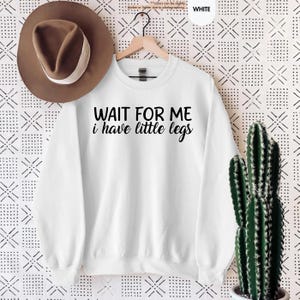 Pode incluir: Uma camisola branca com o texto "WAIT FOR ME i have little legs" em letras pretas. Um chap&eacute;u castanho est&aacute; pendurado acima da camisola. Um cacto num vaso branco est&aacute; no lado direito da imagem.