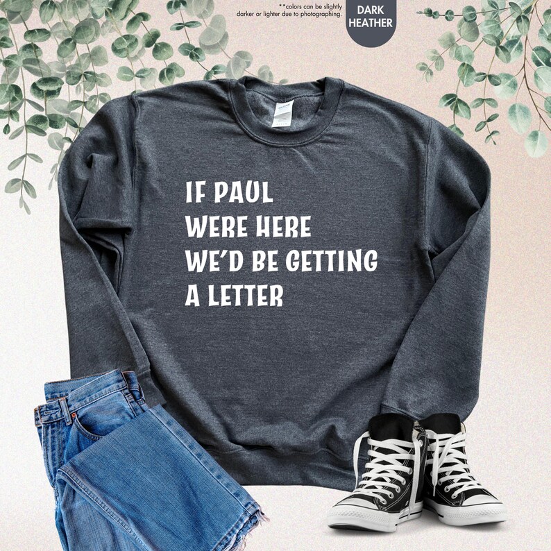 May include: Dark heather gray sweatshirt with the text "IF PAUL WERE HERE WE'D BE GETTING A LETTER" in white. The sweatshirt is paired with blue jeans and black and white high-top sneakers.