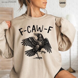 May include: Sand-colored sweatshirt with the text "F-CAW-F" above a detailed black raven illustration with outstretched wings. The sweatshirt is a neutral tone, suitable for various outfits. The design is a graphic print.