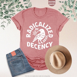May include: Heather mauve t-shirt with the text "RADICALIZED BY BASIC DECENCY" and a dove graphic. The shirt is paired with blue jeans and a tan fedora hat. The image is styled with eucalyptus leaves.