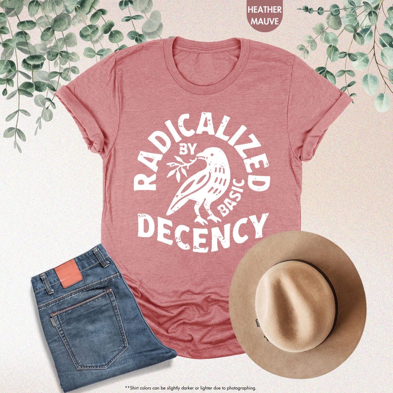 May include: Heather mauve t-shirt with the text "RADICALIZED BY BASIC DECENCY" and a dove graphic. The shirt is paired with blue jeans and a tan fedora hat. The image is styled with eucalyptus leaves.