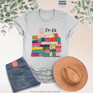May include: Athletic heather gray t-shirt with the text "I'm Ok" and a graphic of a hand emerging from a stack of colorful patchwork quilts. The image also includes blue jeans and a brown hat.