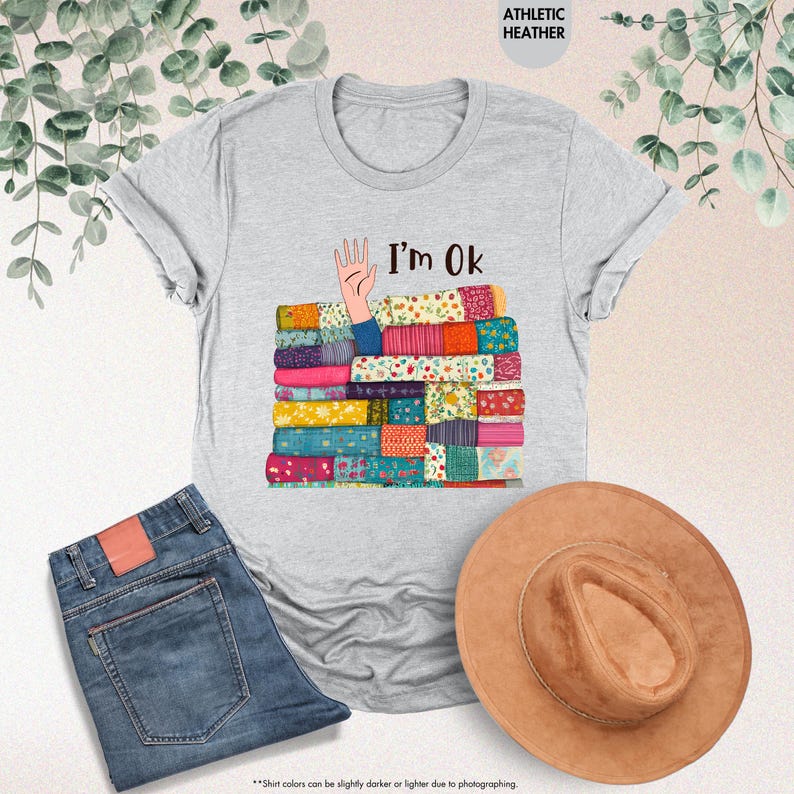 May include: Athletic heather gray t-shirt with the text "I'm Ok" and a graphic of a hand emerging from a stack of colorful patchwork quilts. The image also includes blue jeans and a brown hat.