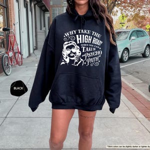 May include: Black hooded sweatshirt with a white graphic of a woman and text that reads "WHY TAKE THE HIGH ROAD? WHEN YOU CAN TAKE THE PSYCHO PATH." The sweatshirt has a front pocket and a drawstring hood. The word "BLACK" is in a black shape.