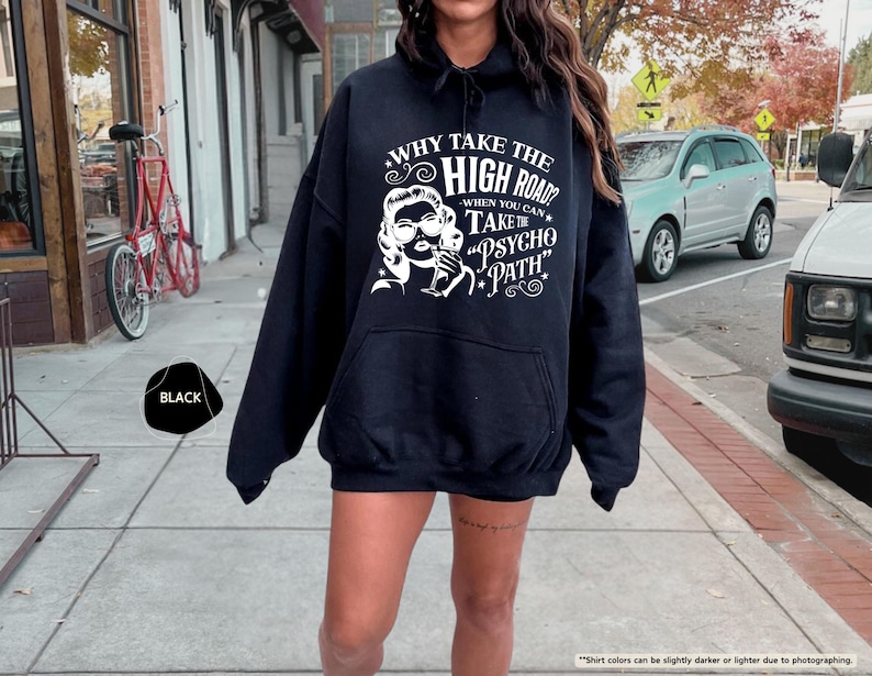 May include: Black hooded sweatshirt with a white graphic of a woman and text that reads "WHY TAKE THE HIGH ROAD? WHEN YOU CAN TAKE THE PSYCHO PATH." The sweatshirt has a front pocket and a drawstring hood. The word "BLACK" is in a black shape.