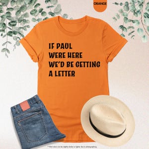 May include: Orange t-shirt with the text "IF PAUL WERE HERE WE'D BE GETTING A LETTER." The shirt is paired with blue jeans and a straw hat. The image also includes a color swatch labeled "ORANGE."