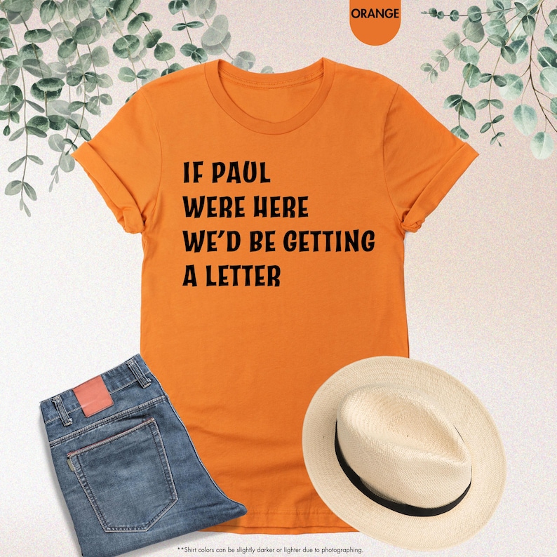 May include: Orange t-shirt with the text "IF PAUL WERE HERE WE'D BE GETTING A LETTER." The shirt is paired with blue jeans and a straw hat. The image also includes a color swatch labeled "ORANGE."