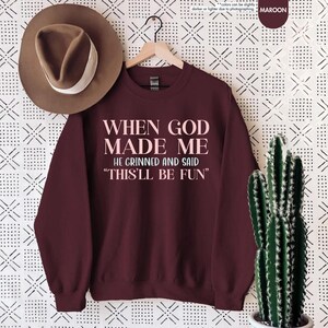 Puede incluir: Sudadera granate con el texto "WHEN GOD MADE ME HE GRINNED AND SAID 'THIS'LL BE FUN'" en rosa claro. Un sombrero marr&oacute;n cuelga encima de la sudadera. Un cactus en una maceta blanca est&aacute; a la derecha.