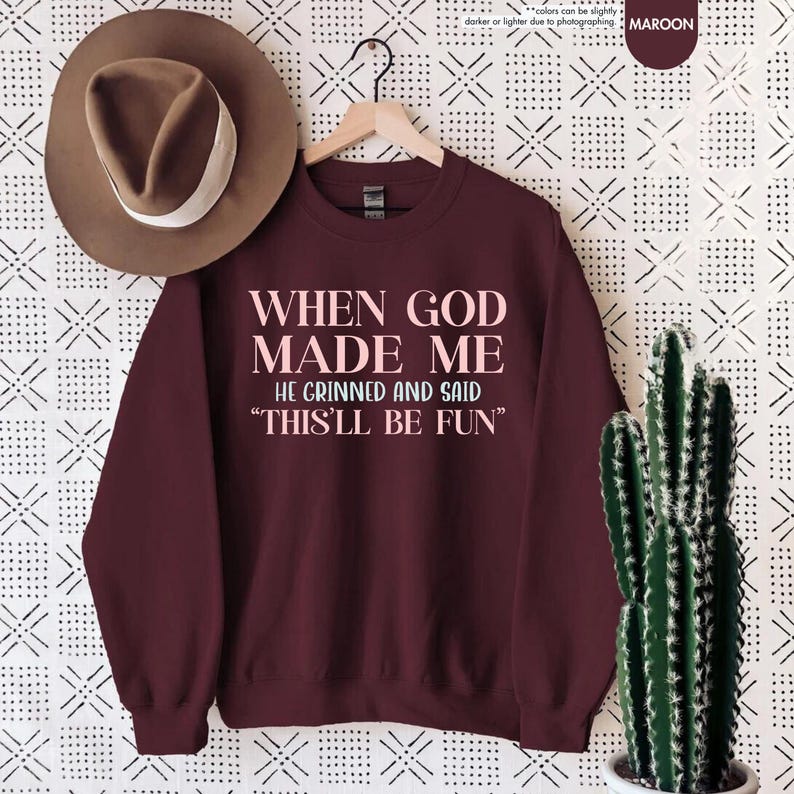 Puede incluir: Sudadera granate con el texto "WHEN GOD MADE ME HE GRINNED AND SAID 'THIS'LL BE FUN'" en rosa claro. Un sombrero marr&oacute;n cuelga encima de la sudadera. Un cactus en una maceta blanca est&aacute; a la derecha.