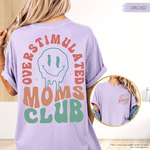 K&ouml;nnte beinhalten: Ein helllila T-Shirt mit dem Schriftzug "OVERSTIMULATED MOMS CLUB" in Retro-Schrift und einem schmelzenden Smiley-Gesicht. Das Shirt hat auch eine kleinere Grafik auf der Vorderseite. Das Shirt hat eine lockere Passform.