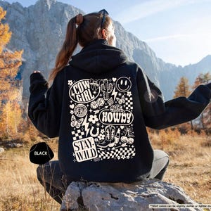 K&ouml;nnte beinhalten: Schwarzer Kapuzenpullover mit einem Grafikdesign auf der R&uuml;ckseite. Das Design enth&auml;lt die W&ouml;rter "Cow Girl", "Howdy" und "Stay Wild" sowie einen Kaktus, Stiefel und ein Smiley. Der Kapuzenpullover wird im Freien gezeigt.