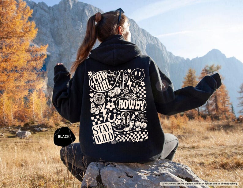 K&ouml;nnte beinhalten: Schwarzer Kapuzenpullover mit einem Grafikdesign auf der R&uuml;ckseite. Das Design enth&auml;lt die W&ouml;rter "Cow Girl", "Howdy" und "Stay Wild" sowie einen Kaktus, Stiefel und ein Smiley. Der Kapuzenpullover wird im Freien gezeigt.