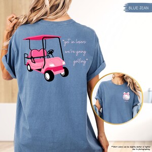 May include: Blue jean t-shirt with a pink golf cart graphic and the text "get in losers we're going golfing." The shirt also has a small golf ball graphic with a pink bow. The shirt is a comfortable, casual style.