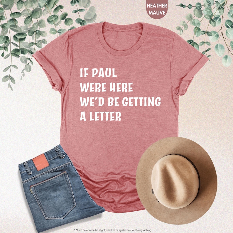 May include: Heather mauve t-shirt with the text "IF PAUL WERE HERE WE'D BE GETTING A LETTER" in white. The image also includes a pair of blue jeans and a tan hat. The shirt is folded, and the jeans are neatly arranged.