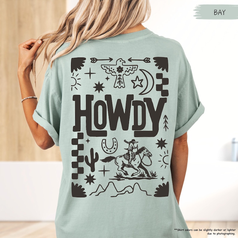 May include: A light blue t-shirt with a black graphic design featuring a cowboy riding a horse, a horseshoe, a cactus, and the word "HOWDY" surrounded by stars and other western-themed symbols.