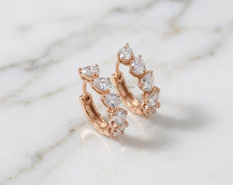 May include: Rose gold hoop earrings adorned with pear-shaped, clear gemstones. The earrings are displayed on a white and gray marble surface. The gemstones are arranged in a repeating pattern around the hoops.