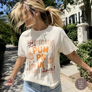 May include: A cream-colored short-sleeve t-shirt featuring a graphic with the text "hey there PUMPKIN Spice!" in orange and brown. The design includes illustrations of a pumpkin, a slice of pie, and a coffee cup, all set against vertical stripes.