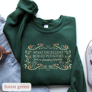 Puede incluir: Sudadera verde bosque con cuello redondo. La parte delantera presenta un dise&ntilde;o decorativo con el texto "WHAT EXCELLENT BOILED POTATOES Such an Exemplary Vegetable Est. 1813". La sudadera est&aacute; sobre una superficie blanca.