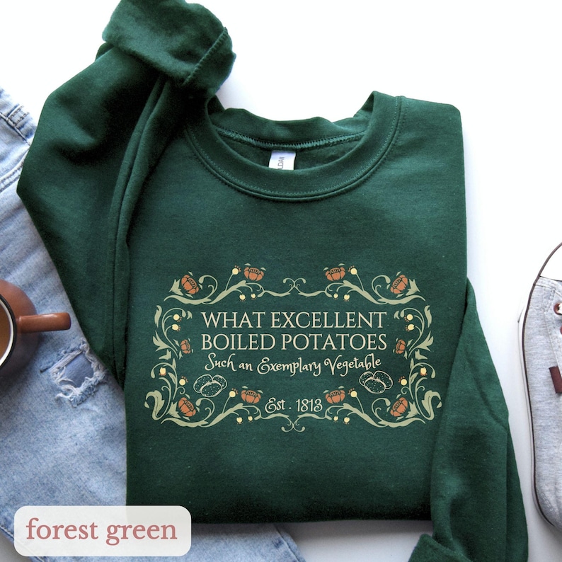 Puede incluir: Sudadera verde bosque con cuello redondo. La parte delantera presenta un dise&ntilde;o decorativo con el texto "WHAT EXCELLENT BOILED POTATOES Such an Exemplary Vegetable Est. 1813". La sudadera est&aacute; sobre una superficie blanca.