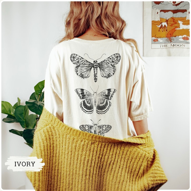 K&ouml;nnte beinhalten: Ein wei&szlig;es T-Shirt mit drei schwarzen und wei&szlig;en Schmetterlingsmotiven auf der R&uuml;ckseite. Das Shirt ist elfenbeinfarben.