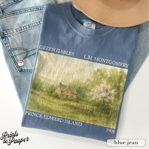 Könnte beinhalten: Ein blaues Comfort Colors T-Shirt mit einem Landschaftsdruck eines Hauses und dem Text "Green Gables, L.M. Montgomery, Prince Edward Island, 1908." Das T-Shirt liegt auf einer blauen Jeans, mit einer goldenen Halskette und einem beigen Hut.