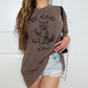 May include: A brown oversized t-shirt with a graphic of a raccoon holding a flower and the text "BE KIND...of feral".