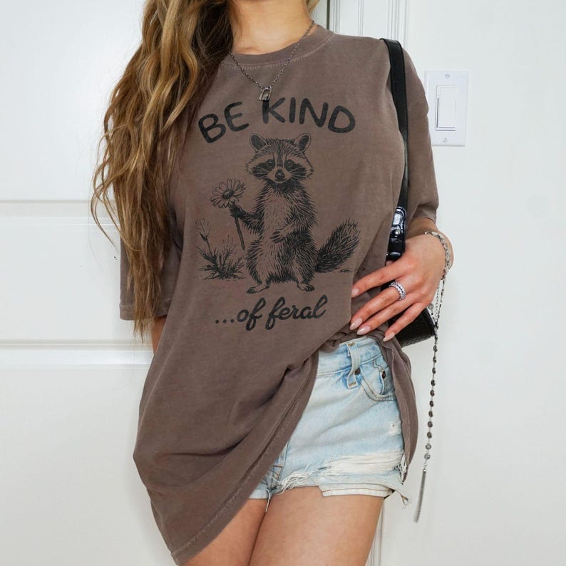May include: A brown oversized t-shirt with a graphic of a raccoon holding a flower and the text "BE KIND...of feral".