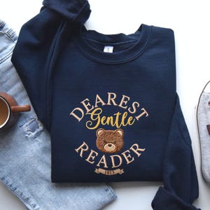 May include: A navy blue sweatshirt with the embroidered text "Dearest Gentle Reader" and a teddy bear graphic. The design is in gold and brown thread. The sweatshirt is displayed with blue jeans and a coffee mug.