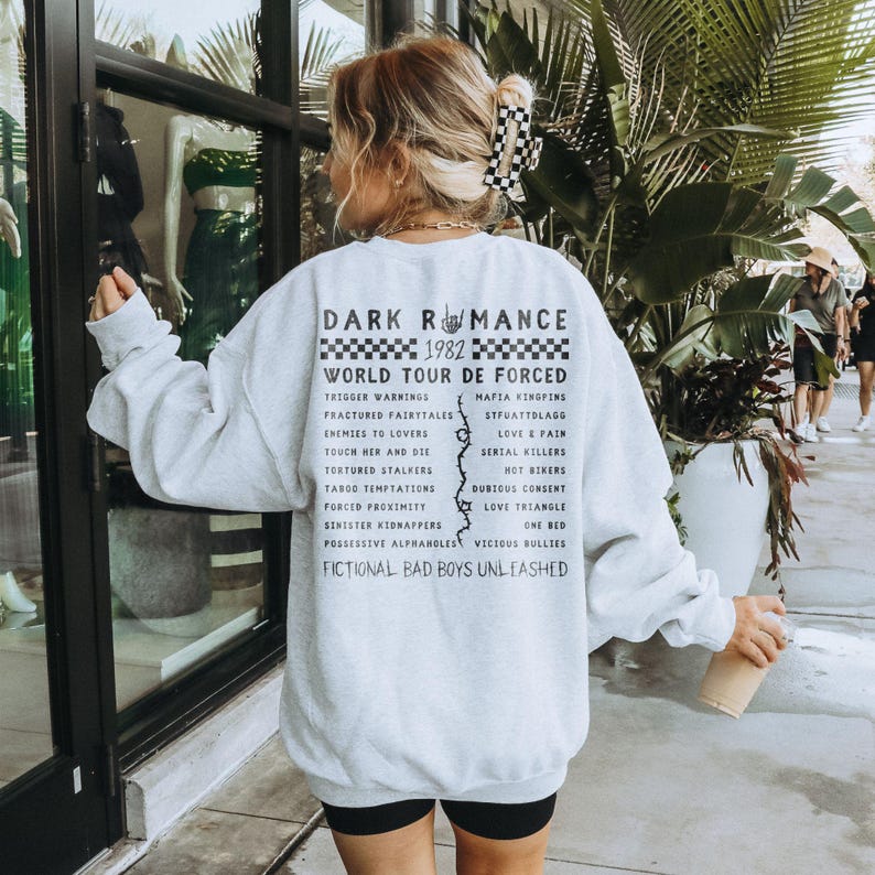 Puede incluir: Una sudadera gris claro con el texto "DARK ROMANCE 1982 WORLD TOUR DE FORCED" y una lista de frases. La persona lleva pantalones cortos negros y tiene el pelo rubio con una pinza para el pelo a cuadros.