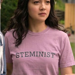 Op de afbeelding: Een T-shirt met korte mouwen en een ronde hals in een gedempte mauve kleur. Het shirt is voorzien van het woord STEMINIST in vetgedrukte, zwarte, schreefloze hoofdletters in het midden van de borst. Het kledingstuk is van effen, ondoorzichtige stof.