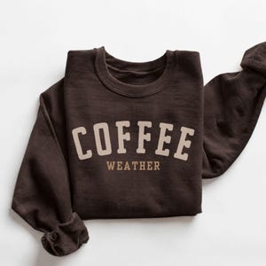 Könnte beinhalten: Ein dunkelbraunes Sweatshirt mit Rundhalsausschnitt, das den Text COFFEE in einer großen, geschwungenen, hellbeigen Schrift zeigt, wobei das Wort WEATHER in einer kleineren, geraden, hellbeigen Schrift direkt unter dem geschwungenen Text steht.
