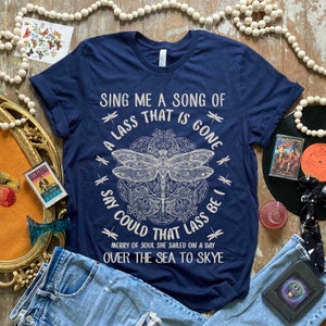 May include: A navy blue t-shirt with a white graphic design featuring a dragonfly and the text "Sing me a song of a lass that is gone, say could that lass be I, merry of soul she sailed on a day over the sea to Skye".