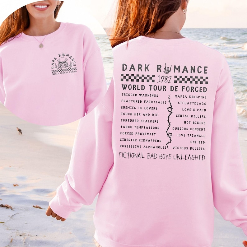 Puede incluir: Una sudadera rosa claro con el texto "DARK ROMANCE" y "WORLD TOUR DE FORCED" impreso en la parte delantera y trasera. La parte trasera presenta una lista de frases y el a&ntilde;o "1982". La sudadera tiene cuello redondo y mangas largas.