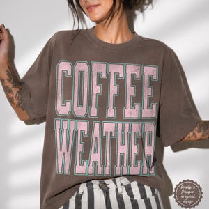 Könnte beinhalten: Ein braunes Kurzarm-T-Shirt mit dem Text COFFEE WEATHER in großen, abgenutzten rosa Blockbuchstaben mit türkisfarbener Umrandung. Das Shirt hat einen Rundhalsausschnitt und eine lockere Passform mit einem Vintage-inspirierten Grafikdesign auf der Vorderseite.