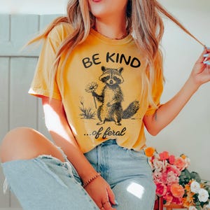 May include: A yellow t-shirt with a black graphic of a raccoon holding a daisy and the text "Be Kind... of feral".