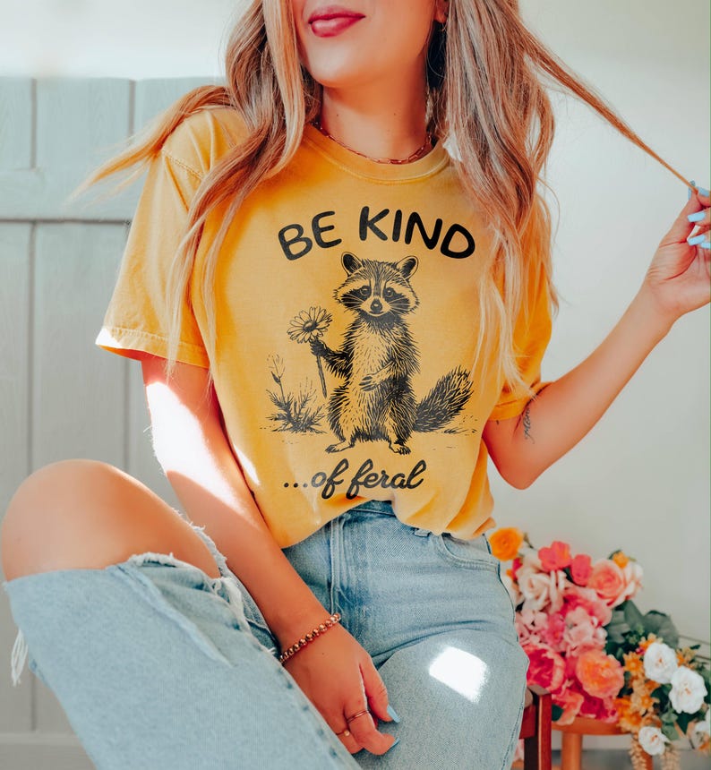 May include: A yellow t-shirt with a black graphic of a raccoon holding a daisy and the text "Be Kind... of feral".