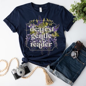 May include: Navy blue t-shirt with the text "Dearest Gentle Reader" in a floral design. The design features purple flowers and green leaves within a white frame. The shirt is paired with denim shorts and sunglasses.