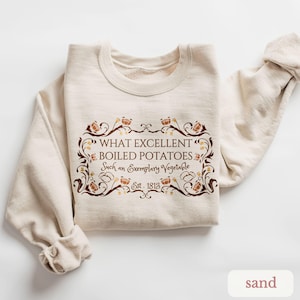 May include: A sand-colored sweatshirt with the text "WHAT EXCELLENT BOILED POTATOES Such an Exemplary Vegetable Est. 1813" in a decorative brown frame. The sweatshirt is folded neatly, with the sleeves visible.