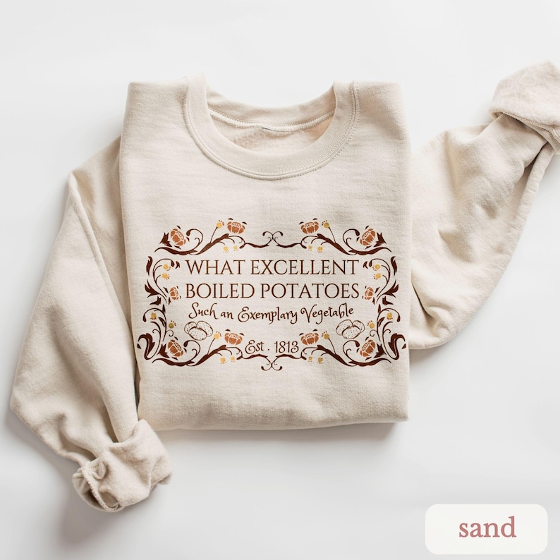 Puede incluir: Una sudadera color arena con el texto "WHAT EXCELLENT BOILED POTATOES Such an Exemplary Vegetable Est. 1813" en un marco decorativo marr&oacute;n. La sudadera est&aacute; doblada, con las mangas visibles.