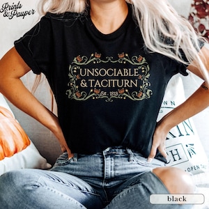 Puede incluir: Camiseta negra con la frase "UNSOCIABLE & TACITURN" en un marco decorativo, con "Est. 1813" debajo. La modelo lleva vaqueros azules.
