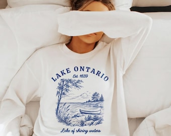 May include: A white long-sleeve crewneck sweatshirt featuring a blue graphic print. The design includes the text Lake Ontario, Est. 1639, and Lake of shining waters, accompanied by an illustration of a canoe on a lake shore with trees.