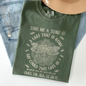 Könnte beinhalten: Ein grünes T-Shirt mit einem weißen grafischen Druck einer Libelle und dem Text "Sing me a song of a lass that is gone, say could that lass be I, merry of soul she sailed on a day over the sea to Skye."
