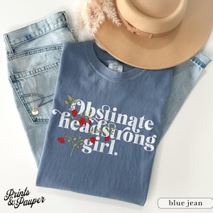 May include: A blue t-shirt with the words "Obstinate & Headstrong Girl" in white, decorated with red roses and green leaves. The shirt is styled with light blue jeans and a tan hat. A gold necklace is visible.