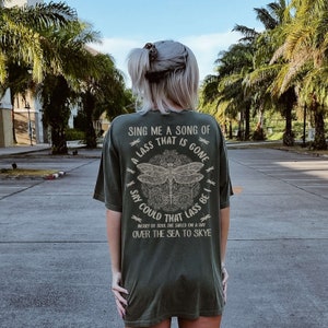 Puede incluir: Una camiseta verde oscuro con un gráfico de libélula y el texto "Sing me a song of a lass that is gone, say could that lass be I, merry of soul she sailed on a day over the sea to Skye".