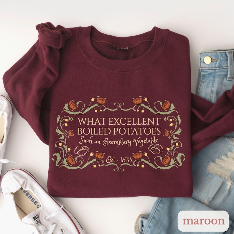 Puede incluir: Sudadera granate con el texto "WHAT EXCELLENT BOILED POTATOES Such an Exemplary Vegetable Est. 1813" en un marco decorativo. El dise&ntilde;o incluye ilustraciones de patatas y hojas. La palabra "maroon" es visible.