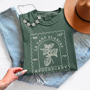 May include: A dark green t-shirt featuring a white graphic with the text "LA DAME BLANCHE APOTHECARY" and a botanical illustration. The shirt is styled with blue jeans, a brown hat, and a floral necklace. The design has a vintage apothecary theme.