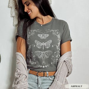 Peut inclure: T-shirt de couleur asphalte avec un motif blanc de trois papillons dans un cadre floral, avec des phases lunaires. Le t-shirt est porté avec un cardigan clair et une ceinture marron. Le mot 'ASPHALT' est visible en bas.