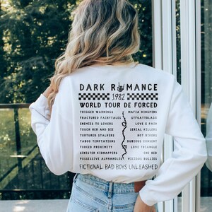 Puede incluir: Sudadera blanca de manga larga con el texto "DARK ROMANCE" y "WORLD TOUR DE FORCED" en negro. La parte trasera de la camisa muestra varias frases y palabras en negro, incluyendo "1982", "Trigger Warnings" y "Fictional Bad Boys Unleashed".