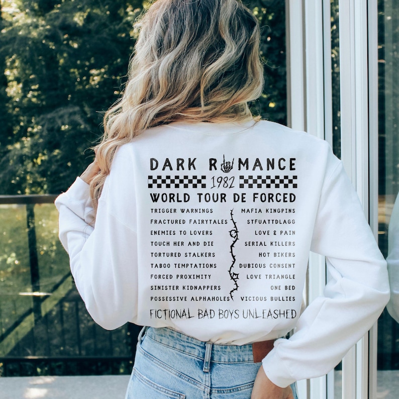 Puede incluir: Sudadera blanca de manga larga con el texto "DARK ROMANCE" y "WORLD TOUR DE FORCED" en negro. La parte trasera de la camisa muestra varias frases y palabras en negro, incluyendo "1982", "Trigger Warnings" y "Fictional Bad Boys Unleashed".