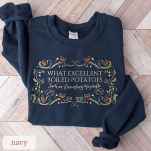 Puede incluir: Sudadera azul marino con el texto "WHAT EXCELLENT BOILED POTATOES Such an Exemplary Vegetable Est. 1813" en un marco decorativo. El dise&ntilde;o incluye ilustraciones de patatas y elementos florales en tonos verdes y naranjas.